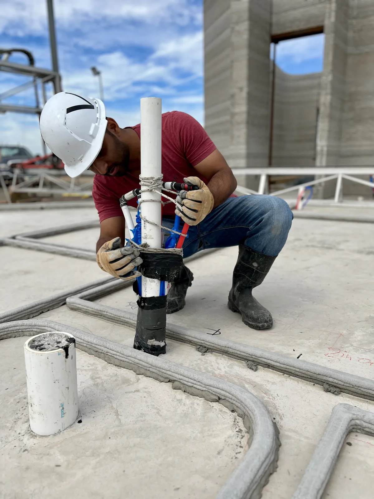 Sebin Joseph in a white hard hat and work gloves, fitting plumbing lines into the first printed layers of a concrete wall on site