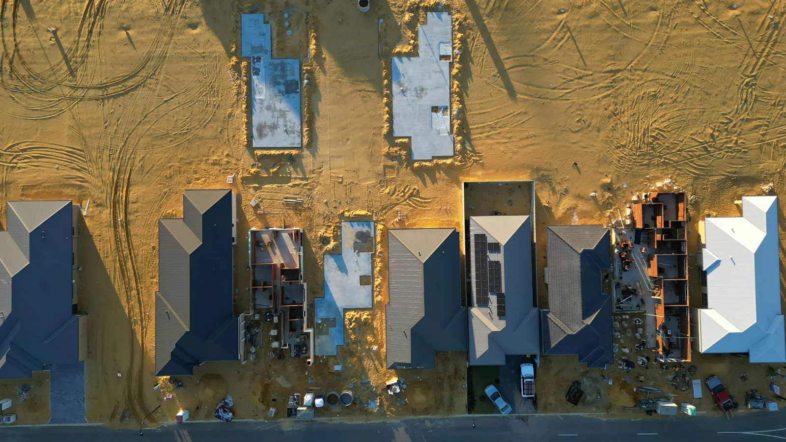 An aerial view of one street of new homes at every stage at once: bare slabs, walls going up and finished roofs