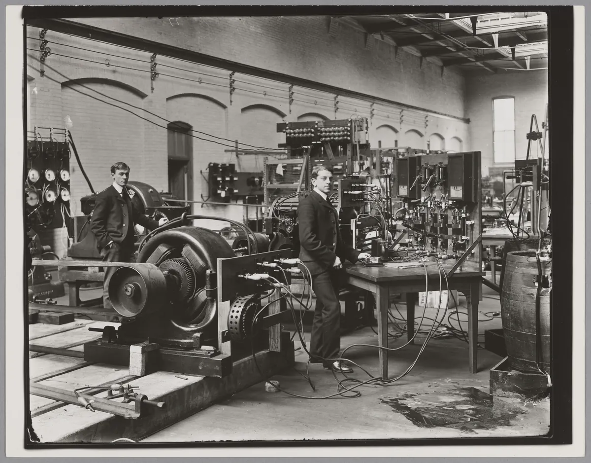 Two men in suits among dynamos, switchboards and cables in an electrical engineering laboratory in the 1890s