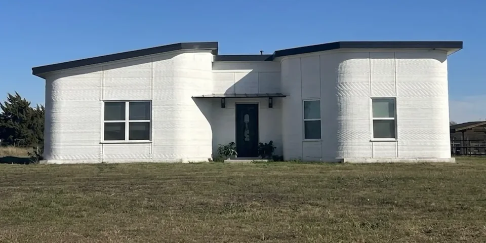 The house finished: a single-story house with white printed concrete walls, rounded corners and a dark roof, on a grass lot under a clear sky.
