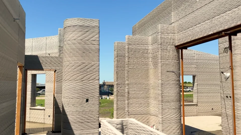 Inside the house before the roof: printed concrete walls at full height, a round printed column, and window and door openings under steel lintels looking out to the street.